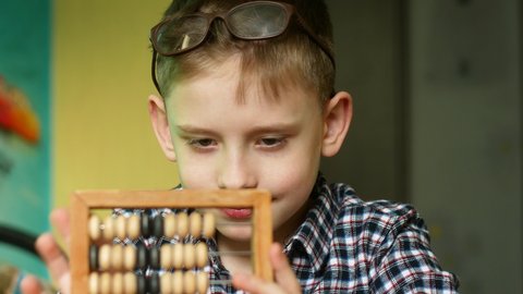Caucasian boy 7-8 years old in glasses sitting at the table at home considers abacus wooden. Child learns arithmetic using old abacus. Education theme at home. Selective focus