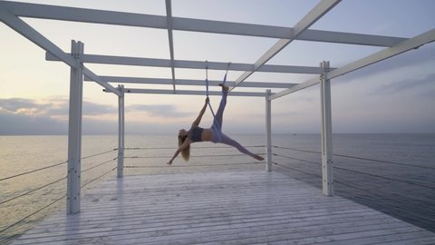 Young blonde woman in modern tracksuit does yoga exercises hanging on special slings fastened to wooden planks on sea empty pier against cloudy sky