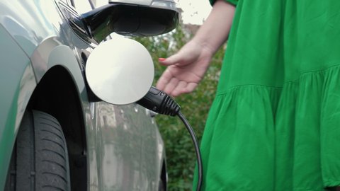Female hands opens charging port lid an electric car, plugging in power supply cable to charge EV. Unrecognizable woman in green dress replenishes energy of vehicle on background of house and trees.