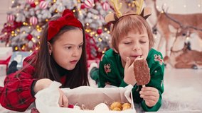 4k video portrait of two cute little children eating Christmas sweets. Craft chocolate handmade sweets in hands of kids. Brother eating sweets near christmas tree.