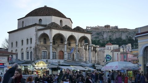 ATHENS - JANUARY 19, 2018: Young man and woman smiling in people crowd on Monastiraki Square in Athens at the evening, the view on Tzistarakis Mosque and the Parthenon. Medium shot.
