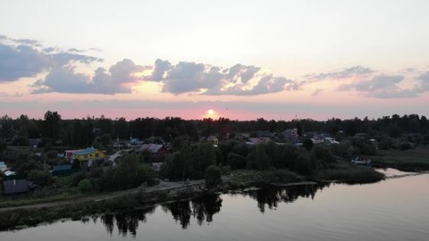Sunset at river town, small houses seen down, aerial camera move up, reveal panoramic view of Novaya Ladoga. Setting sun seen at horizon line, some clouds on neat colored sky