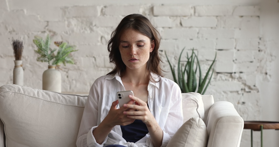 Teenager young woman sit on sofa holds smart phone looks at gadget screen read bad news by sms feels concerned. Concept of damaged cellphone, slow internet connection, need device repair due malware Royalty-Free Stock Footage #1083762262