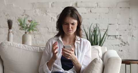 Teenager young woman sit on sofa holds smart phone looks at gadget screen read bad news by sms feels concerned. Concept of damaged cellphone, slow internet connection, need device repair due malware