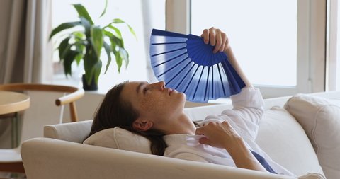 Young freckled woman lean on sofa cushions waving fan, cooling herself at hot day, reduces heat inside living room without air-condition, climate control system. Discomfort, hormonal imbalance concept