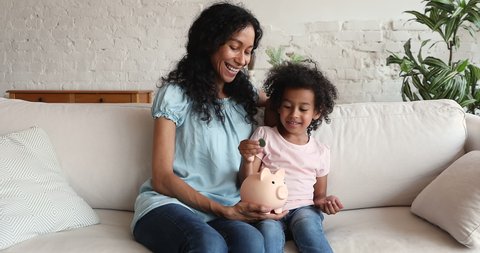 African little 5s cute girl sit on sofa with loving mother counts and drops coins into piggy bank. Save up money for future, tomorrow expenses. Caring parent teach preschool child to be frugal concept