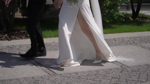 Bride and groom walk along the pavement sidewalk. Legs of man and woman, wedding couple. Girl in a white long dress and veil with flowers bouquet. Male in black suit.