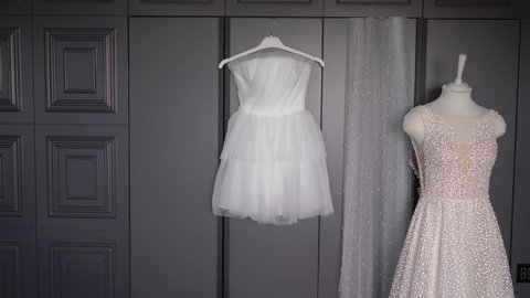 Two wedding dresses, white bridal gown hanging on hanger in bedroom. Morning of the bride. Long and short, white and pink. Bride veil in store.