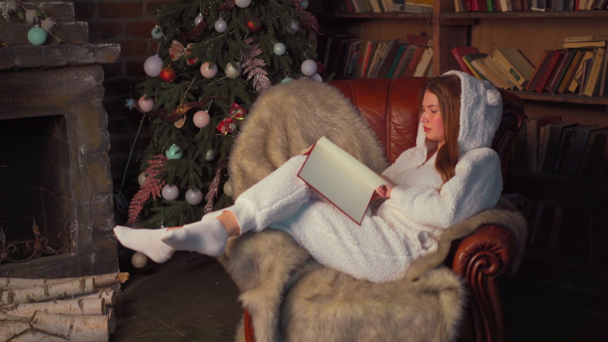 Christmas. Woman reading book sitting cozy leather armchair with Christmas tree in background . Beautiful young caucasian female resting home reads book dressed warm pajamas jumpsuit