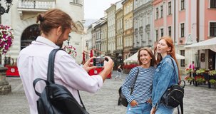 Caucasian friends tourists walking and taking photos on smartphone. Young guy filming shooting pretty women hugging and laughing for camera. Technology, travel, friendship concept.
