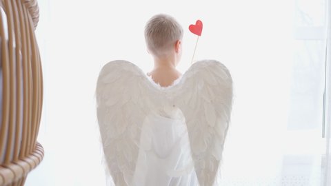 Cupid with white wings and an arrow with a red heart. Valentine's day child dressed as an angel with wings. 4k video in a white studio. Congratulations on Valentines Day. Boy waving his heart