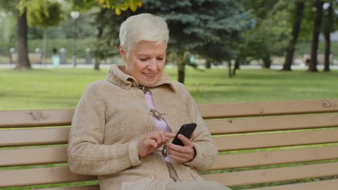 Overjoyed gray-haired mature woman sitting outdoor, using phone browsing smartphone apps, looking at screen, senior female chatting with relatives online, shopping, having fun with mobile device