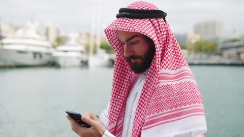 Traditional dressed arab businessman with smartphone in port, reading internet news on mobile phone 