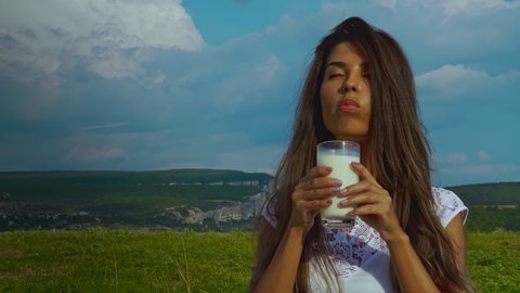A beautiful girl drinks milk from a glass against the backdrop of a green field and mountains. Portrait of a contented woman drinking milk. Attractive lady enjoying a glass of milk in nature