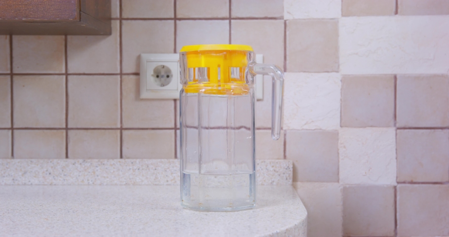 Woman takes glass water filter with yellow cap standing on white marble kitchen table against beige tile wall AT home closeup