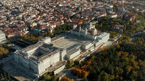 Aerial view of large historic Royal Palace complex at golden hour. Colourful foliage on trees in park of gardens. Tilt up revealing cityscape.