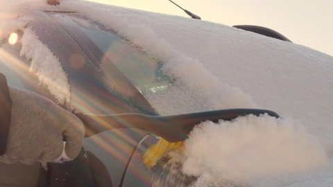 The windshield is covered with a thick layer of snow, which is brushed off with a car brush. A woman cleans snow and ice from her frozen car before driving. Car enthusiasts in the Nordic countries.