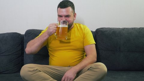 Young man is sitting on gray sofa at home in front of the TV, rejoicing and drinking beer from glass mug. He drinks beer in big gulps and smiles broadly. Relax at home, watch TV. Alcohol