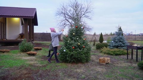 A beautiful and cheerful family with a dog is now decorating a Christmas tree near a country house in winter. The slender mother with two daughters decorates the Christmas tree with toys and balls.