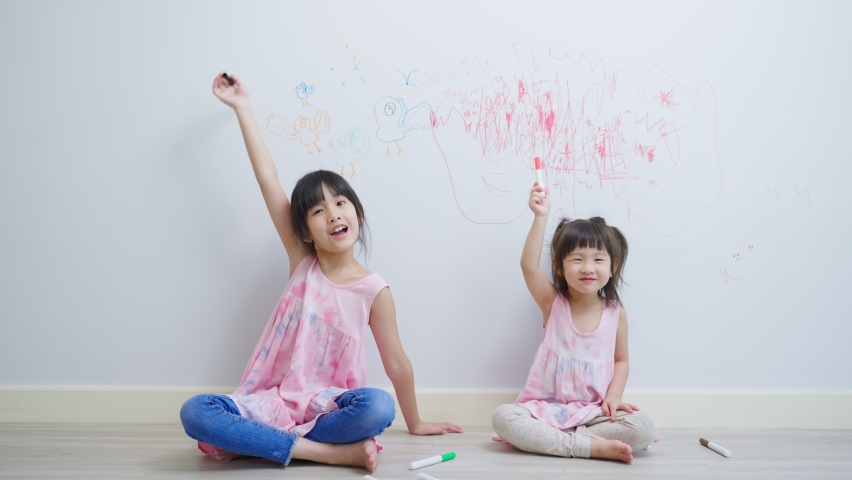 Asian young sibling kid girl enjoy paint on white wall in living room. Little adorable children having fun drawing and coloring art picture with hapiness enjoy creativity at home and looking at camera