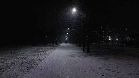 Pedestrian alley at night during snowfall in winter. City lights. Urban lighting with lampposts lantern. Electric energy and electricity power. Residential multi-storey buildings in the background
