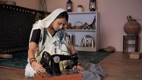A beautiful Indian village woman making clothes on her old hand sewing machine - Employment in Rural India. A rural housewife wearing a traditional saree stitching clothes while sitting on the floo...