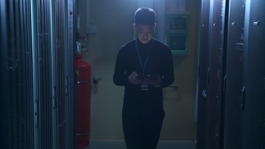 Male engineer working with tablet and inspecting equipment, walking in data office spbas. Front view of young man holds device in hand and examines hardware racks, does analytical work and walks in