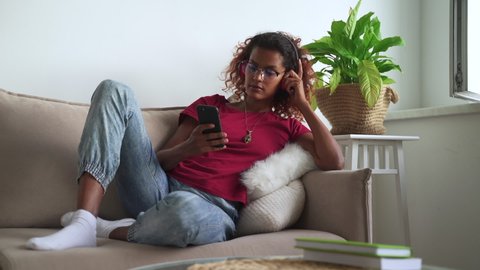 Podcast entertainment show. Pretty young African-American female student with headphones and spbas cellphone chooses radio channel on sofa at home