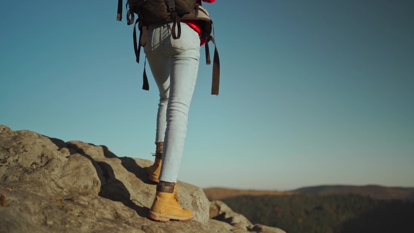 slow motion rear view Young adventurous woman hiker backpacker in a red insulated jacket and jeans hiking up a rocky mountain at sunset above the woods. womn raisinh hands on top of cliff mountain.