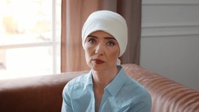 Cancer Concept. Female Wearing Headscarf Looking At Camera During Chemotherapy And Oncology Treatment Sitting On Sofa In Modern Clinic Indoors. Slow Motion