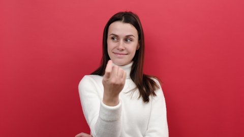 Portrait of young caucasian woman looking playfully and calling with one finger, making beckoning gesture, inviting to come, wears white knitted sweater, posing isolated over red color background wall