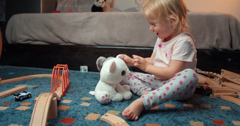 Carefree cute toddler girl is sitting, smiling and playing with stuffed animal toys meanwhile her brother plays with wooden railway road. Playful childhood of children on evening in living room