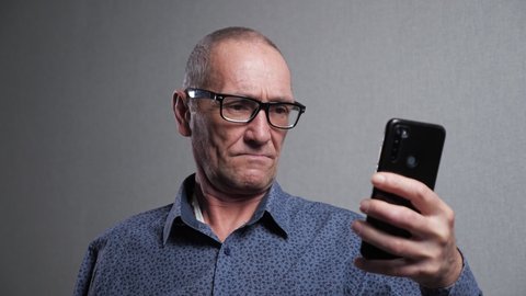 Senior man pensioner with bad eyesight in elegant black-rimmed glasses tries to read message in modern smartphone on grey background closeup.