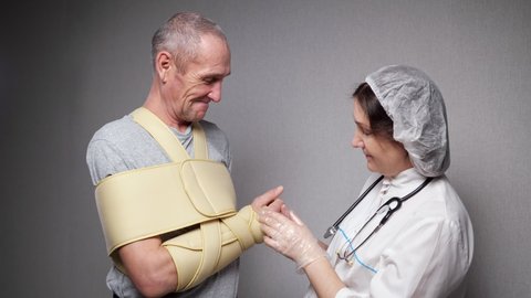 Professional lady orthopedist in uniform checks finger bones and joints workability of elderly man with fixating bandage over torso at grey wall closeup.