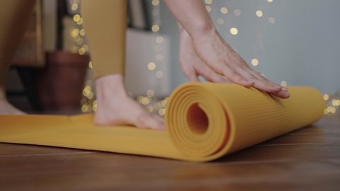 Young barefoot woman wearing sporty yellow leggings unrolls bright-colored yoga mat before practicing against illuminating garland closeup