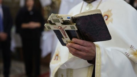 An Orthodox priest holds a cross and a bible in his hands. A priest with a cross in his hands reads a prayer while standing in the church in front of the parishioners.
