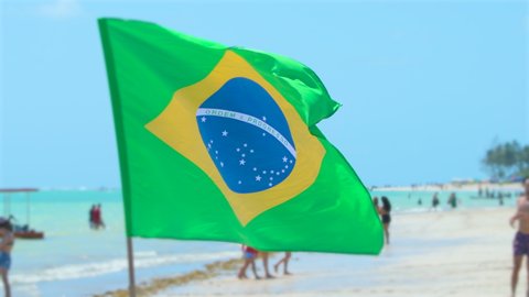 Brazilian flag waving on a beautiful beach of the brazilian northeast. People playing soccer, bathers enjoying the sunny day and the sea on the background. Brazilian tourist destination.