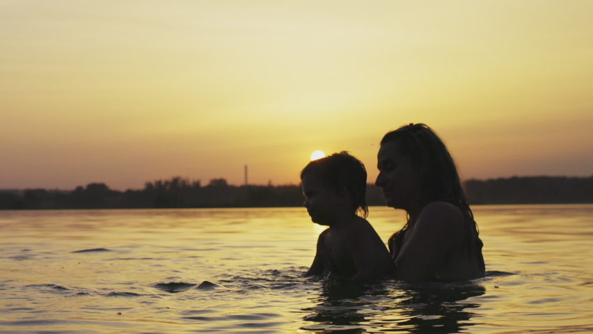 Happy mother with naked laughing baby she holds in arms playing with daughter and splashing in transparent clear blue lake against background of bright sunny summer sunset. HD video slow-motion