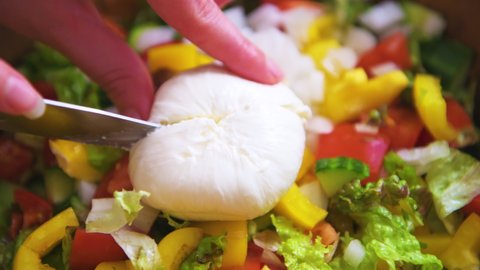 Macro closeup of hand knife person cutting burrata mozzarella Italian cheese ball with creamy center inside on fresh caprese salad with yellow bell peppers, tomatoes crumbled