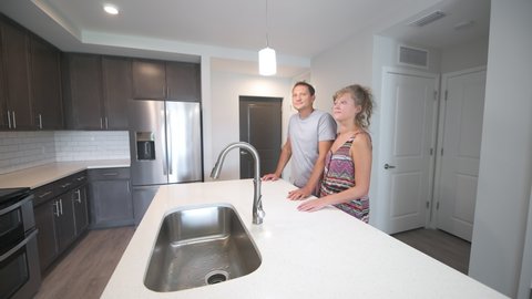 Young couple man woman entering through front door, inspecting empty apartment kitchen island room for sale or rent by stainless steel appliances, refrigerator and sink in modern condo home