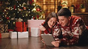 Couple lying at house hall decorated with xmas tree glowing garland texting sms with congrats Merry Christmas relatives and friends using tablet. Buy online, sale, modern tech overuse concept - Powered by Shutterstock - Get 15% off with code: PIKWIZARD15