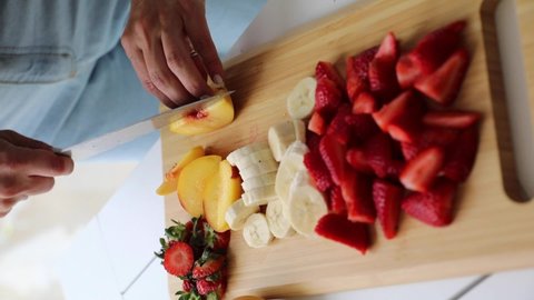 Chopping slicing fresh fruit pear banana peach strawberry with knife in wooden board