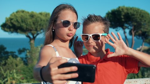 Cheerful children in sunglasses enjoying family weekend at sunny seaside. Smiling teenagers taking selfie on cellphone at sea resort outdoor. Joyful boy and girl grimacing at mobile phone camera