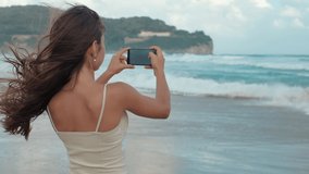 Cheerful girl making video call with mobile phone at sea beach. Joyful teenager cellphone spending summer holiday at seashore. Happy female person filming video at smartphone at wind coastline.