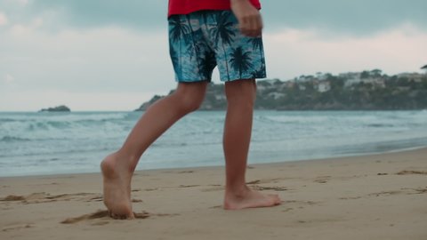 Unknown man legs walking along sea coastline in cloudy day. Unrecognizable boy leaving footprints at wet sand beach seashore. Teen male spending summer holiday at seaside.