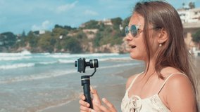 Happy young woman recording video blog at seaside. Cheerful vlogger girl talking to camera at seashore in summer day. Joyful teen female person filming with camera stabilizer outdoors.