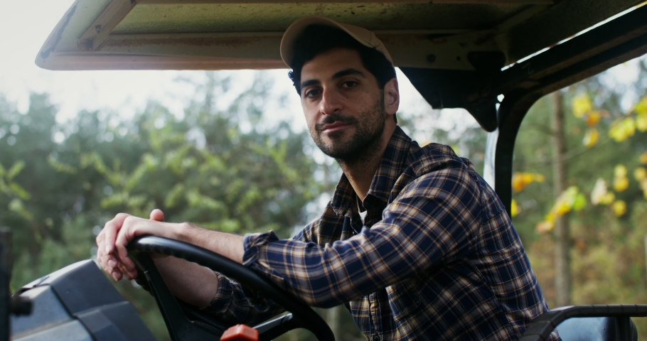 Young black-haired man of European appearance with a beard smiling, while looking at the camera while sitting at the wheel of an agricultural car with a canopy