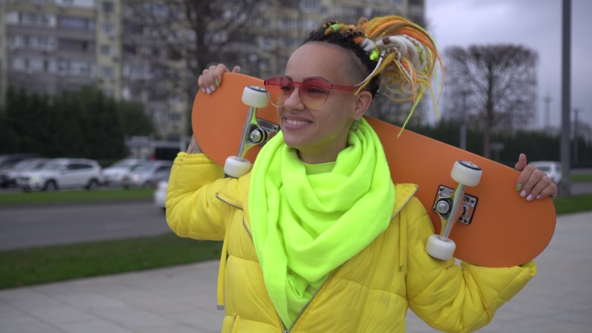 A young happy woman with a bright appearance walks through the city. Stylish modern woman with skateboard outdoors. Youth subculture and lifestyle