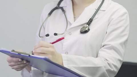 Close up of a doctor a woman in a medical gown with a stethoscope around her neck holding a tablet in her hands and taking notes