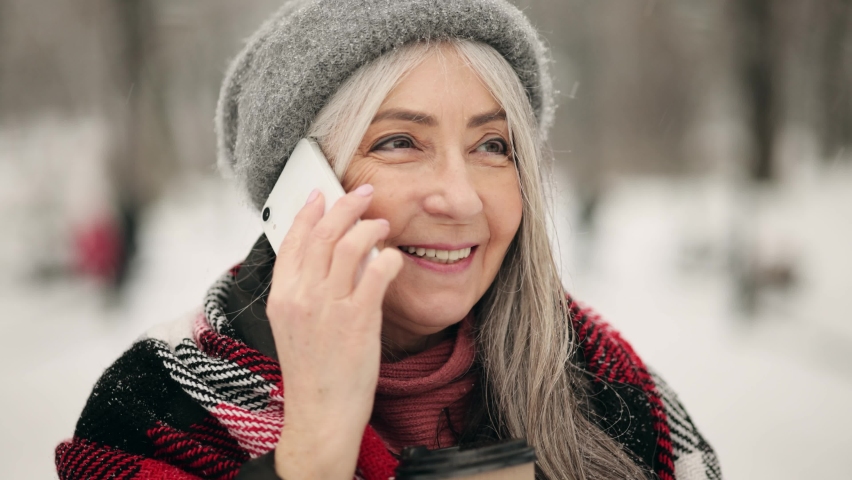 Attractive grey-haired senior woman talking using smartphone in snowy holiday city. Happy smiling middle aged woman walking down the street. Snowing outdoors.